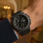 A black dual-display sports watch on a wrist, with a cozy indoor background.